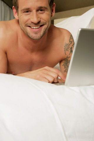 Young unshaven man lying on bed with a laptop (part of), close-up