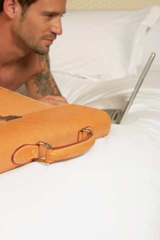 Man lying on bed, using laptop, briefcase in foreground